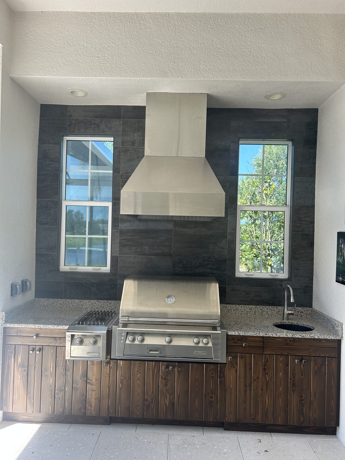Outdoor kitchen with large-format dark porcelain tile wall behind a built-in grill and vent hood